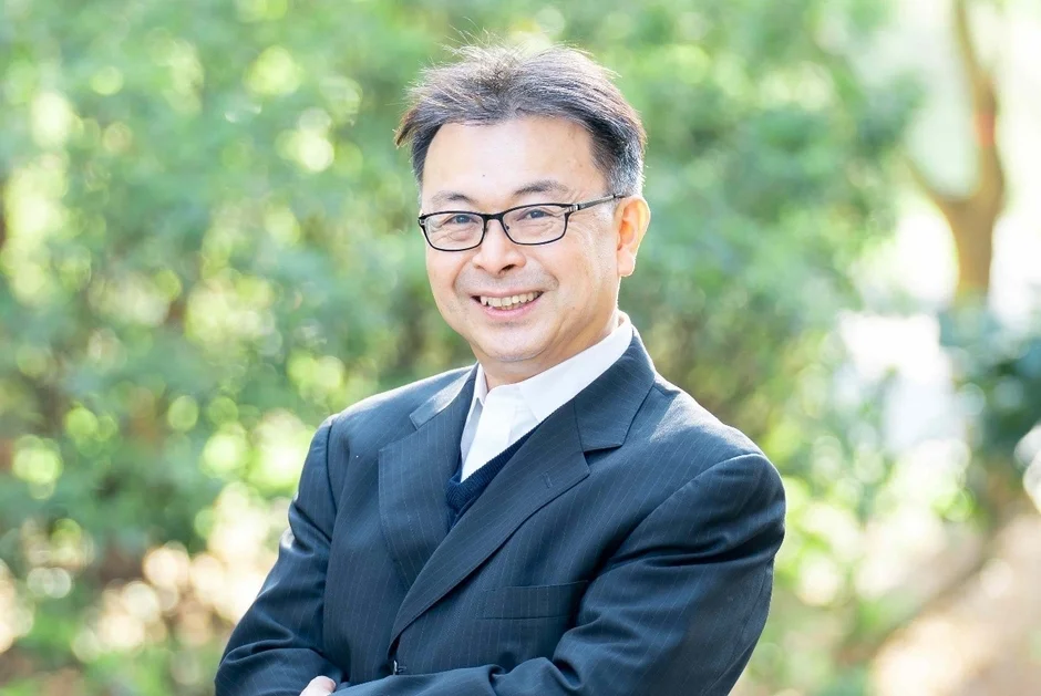 緑豊かな背景を背に、眼鏡をかけた中年の男性がスーツ姿で笑顔を見せているポートレート写真です。