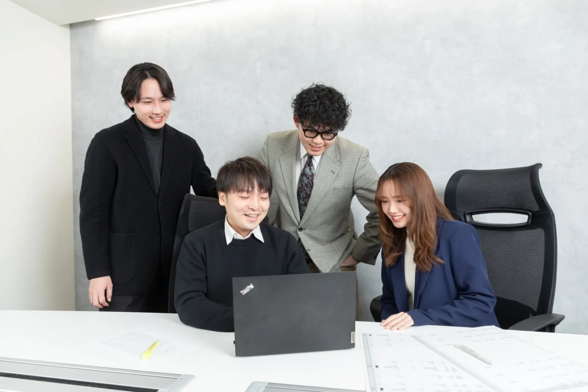 オフィスで4人の若いビジネスパーソンがパソコンを囲んで笑顔で話し合っています。