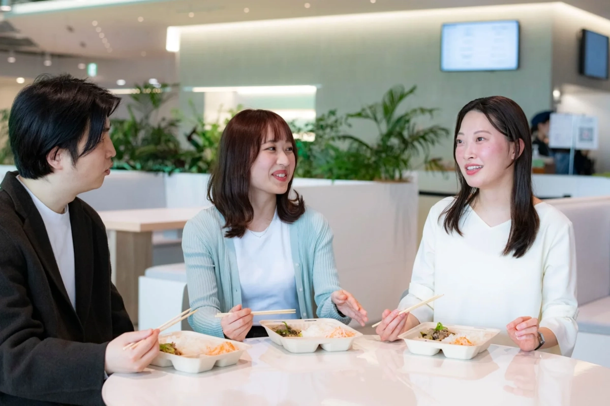 明るくモダンなカフェテリアで、3人の若者が各自の食事をしながら談笑しています。ランチタイムの休憩やミーティング中の様子を捉えた一枚です。