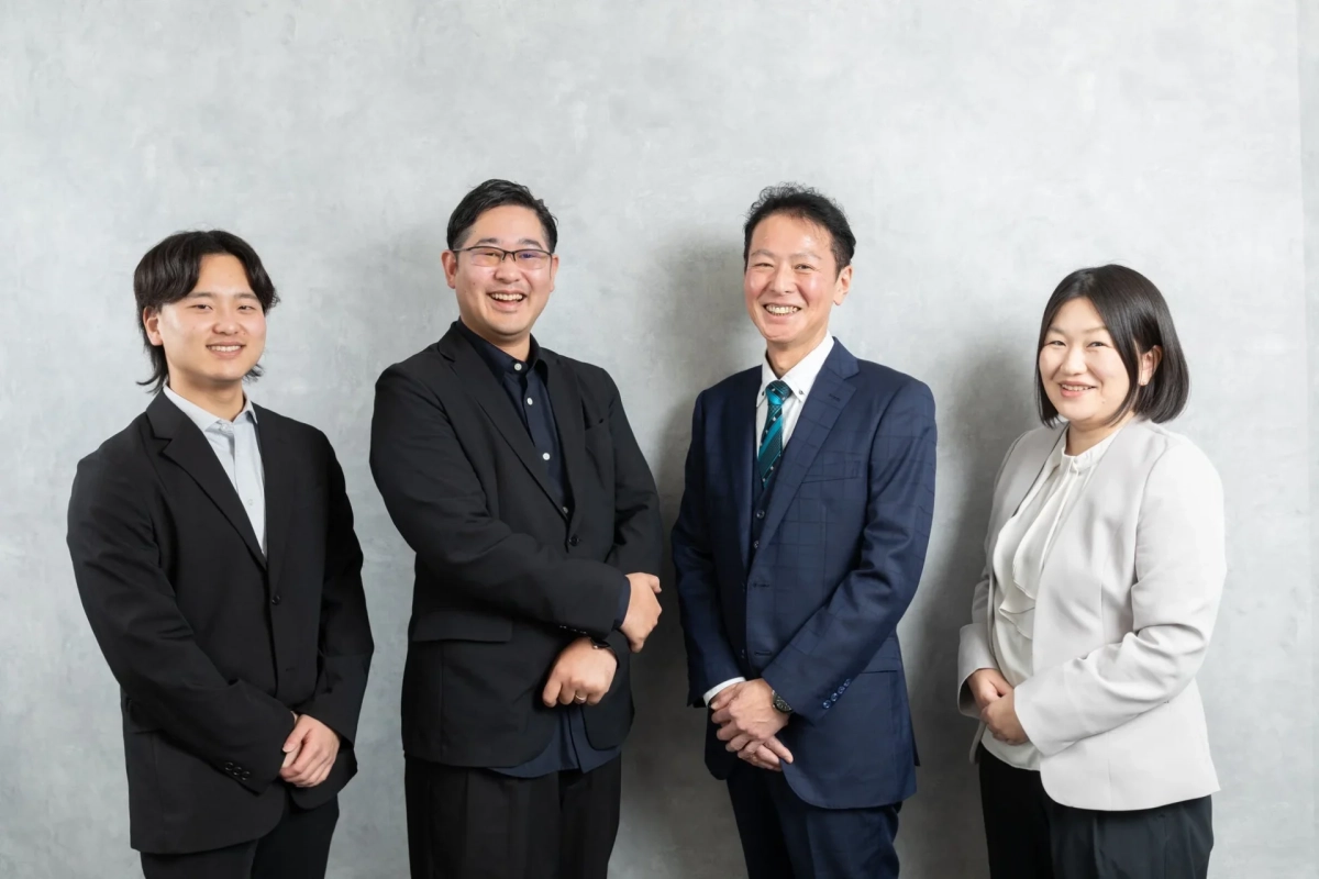 4人のビジネスパーソンが笑顔で並んで立っている集合写真です。