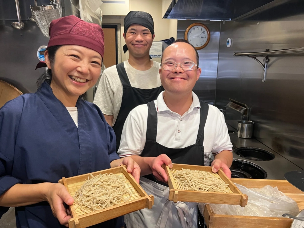 厨房で3人が笑顔でそばを持つ