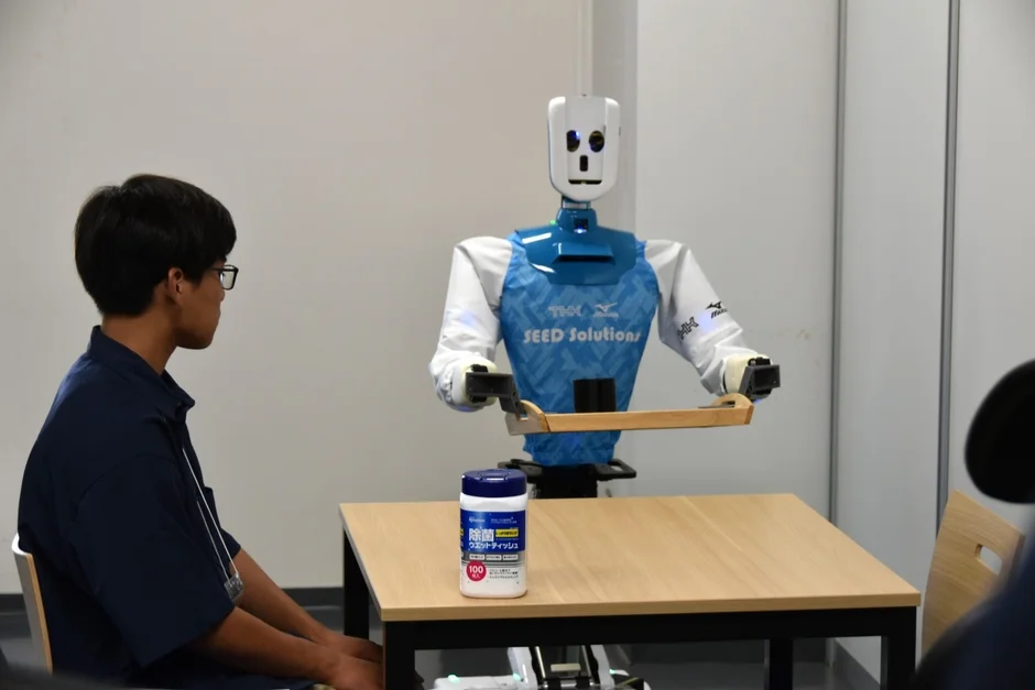 テーブルを挟んで人物とヒューマノイドロボットが向かい合っている。ロボットはトレイを持ち、テーブルには除菌ウェットティッシュが置かれている。サービスロボットの活用やデモンストレーションの場面を示す一枚。
