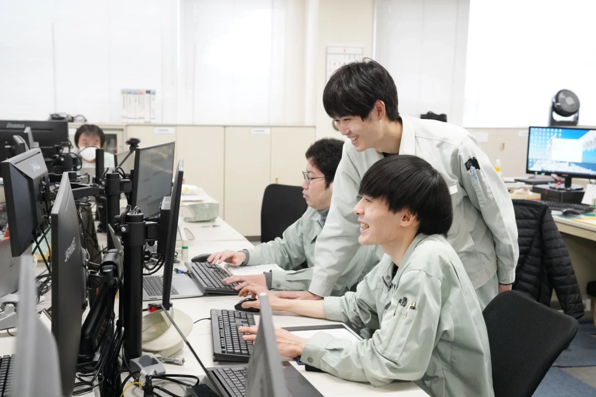 複数の若い男性社員がオフィスでPCに向かって作業しており、一人が立って他の社員と笑顔でコミュニケーションを取っている様子。共同作業や指導の場面を思わせる。