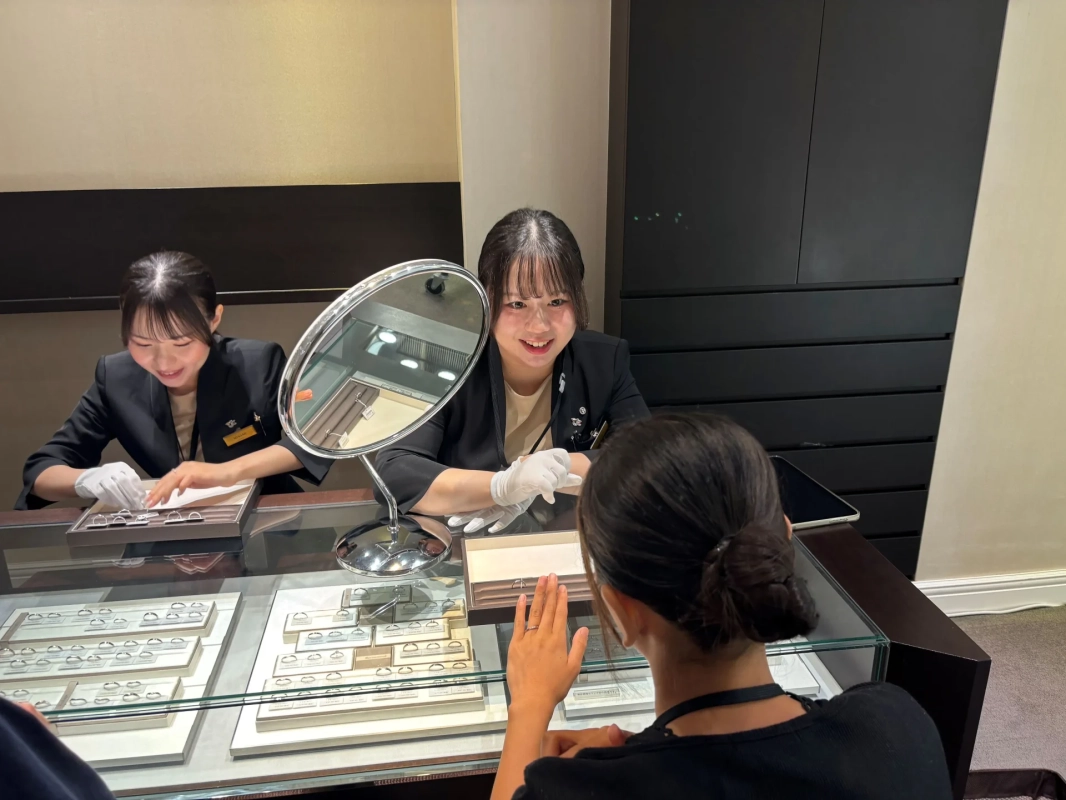 宝石店で二人の女性店員がお客様に指輪を案内している様子です。一人の店員は笑顔で接客し、もう一人はディスプレイを整えています。