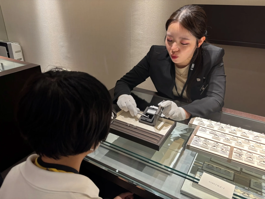 宝石店で女性店員が白い手袋を着用し、顧客にダイヤモンドや指輪を説明している場面です。ガラスケースには多数の指輪が並べられており、真剣な雰囲気で商品選びが行われています。