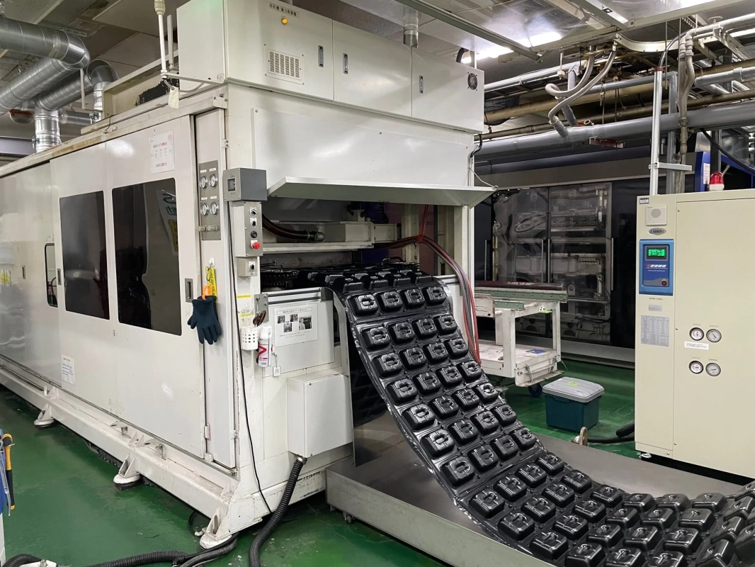 工場内で稼働中の大型成形機が写っています。機械からは黒いプラスチック製の部品が連続して排出されており、製造工程の一部を示しています。多くの配管や制御盤があり、自動化された産業生産の様子がうかがえます。