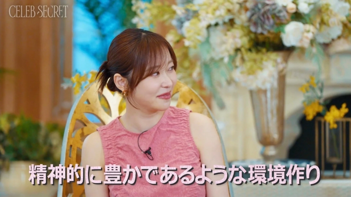 ピンクのトップスを着た女性が、華やかな花飾りのあるスタジオでインタビューを受けている様子