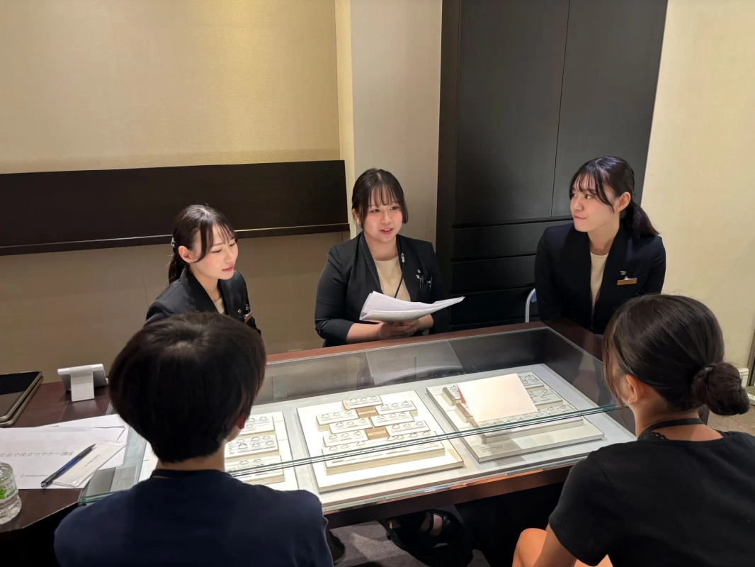 ジュエリーショップらしき店舗で、3人の女性スタッフが2人の顧客に対応している様子です。ガラスケースに並べられた指輪を見ながら、スタッフが書類を使って説明しているようです。マナー講座に関する資料も見受けられます。