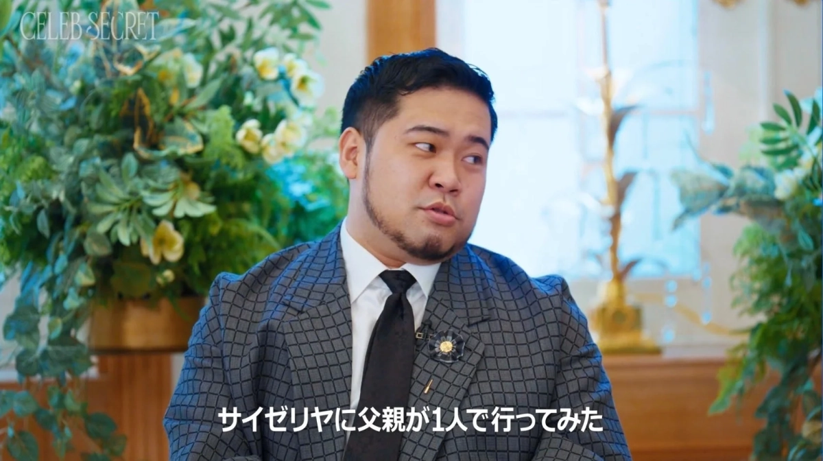 スーツを着た男性が、花が飾られた室内で横を向いて話している様子
