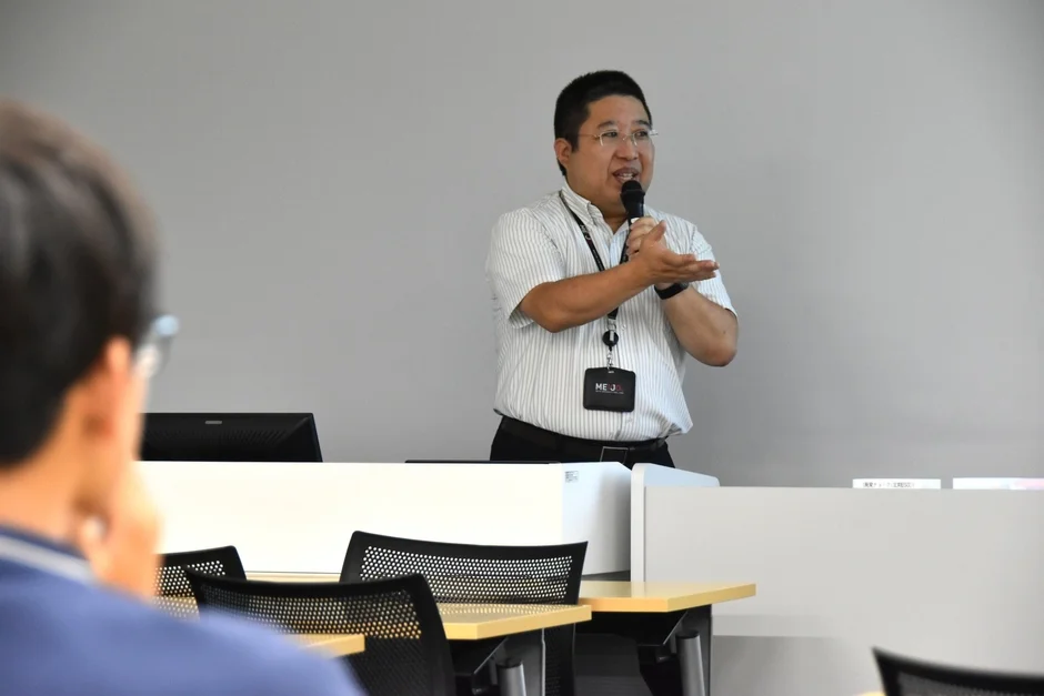 会議室で男性がマイクを持ってプレゼンテーションを行っており、聴講者が熱心に耳を傾けている様子が捉えられています。ビジネスや教育の場面を思わせる一枚です。