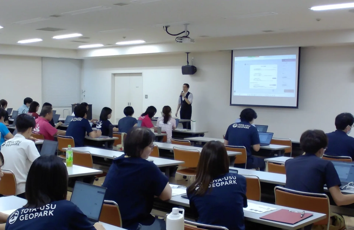 教室で講師がプロジェクターを使い説明しており、参加者は各自のノートパソコンで作業をしています。多くの参加者が「TOYA-USU GEOPARK」と書かれたシャツを着用しています。