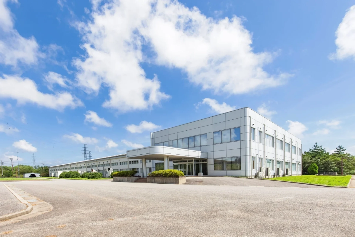 青い空と白い雲が広がる晴れた日に、広大な敷地に建つモダンな白いオフィスビルまたは工場。手前には広い駐車スペースがあり、奥には送電線が見える。
