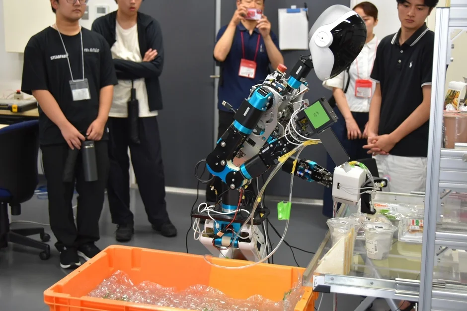 研究室らしき場所で、数人の学生や関係者が見守る中、ロボットアームがオレンジ色の容器の上で作業を行っています。棚には包装された食品のようなものが見え、ロボットの動作を観察している様子が伺えます。