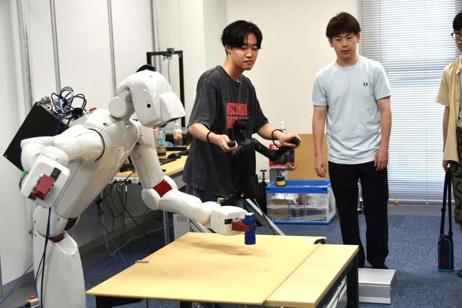 研究室で学生が白いヒューマノイドロボットを操作している様子。卓上の物体を動かすロボットを、別の学生が見守っている。