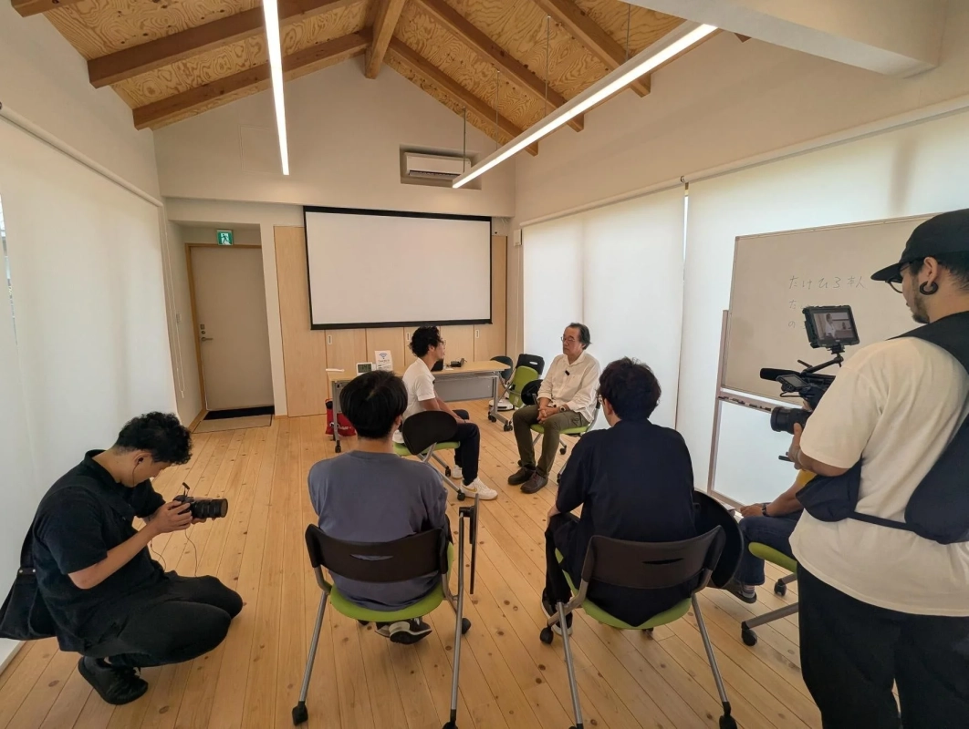 木目調の天井が特徴的な明るい部屋で、数人の男性が集まり、インタビューや撮影が行われている様子です。二人の男性が向かい合って座り、他の人々がカメラでその様子を記録しています。背後には大きなプロジェクタースクリーンとホワイトボードが見えます。