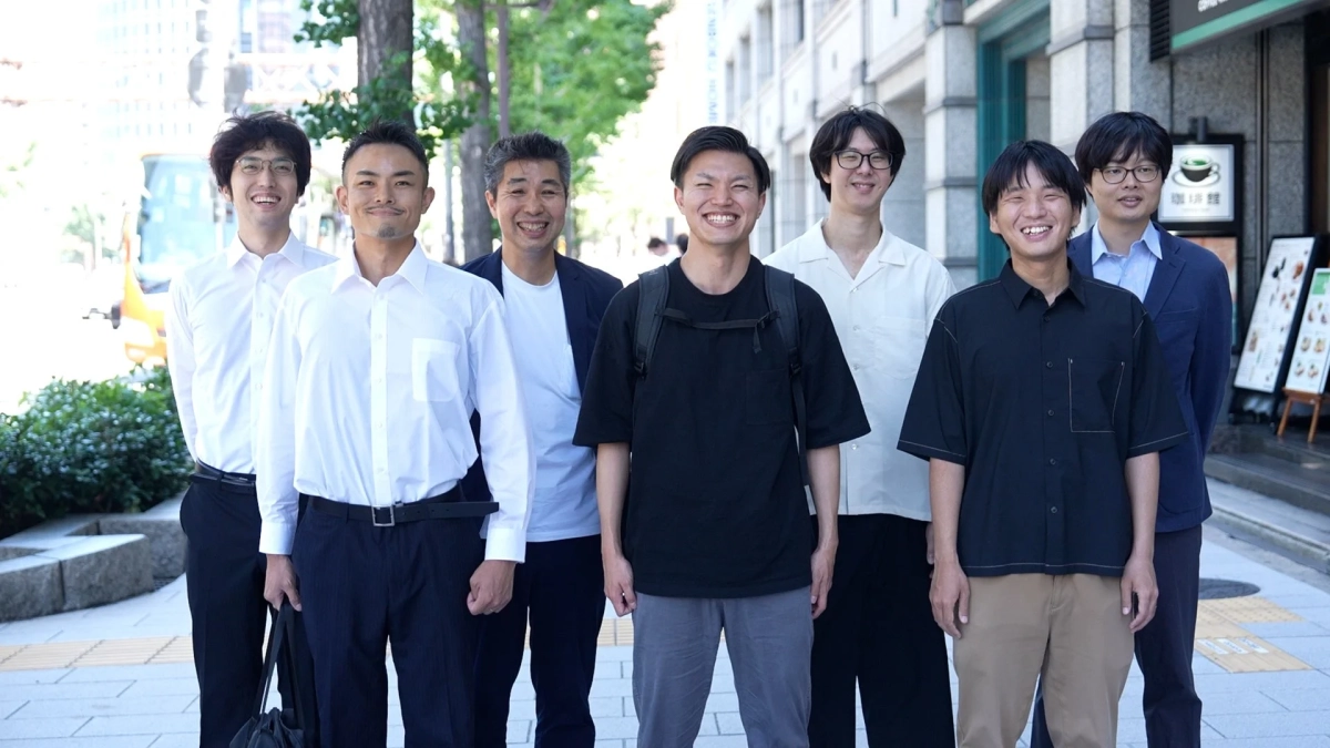 7人の日本人男性が街中の歩道に並んで笑顔で立っている集合写真