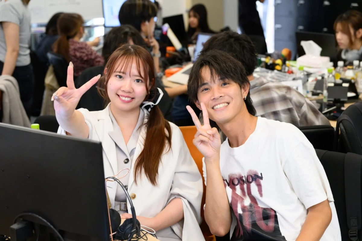 オフィスのような場所で、笑顔の若い男女がカメラに向かってピースサインをしている様子