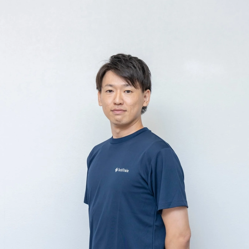 TechTrainのロゴ入り紺色のTシャツを着た男性が、白い背景の前でカメラ目線で立っているポートレート写真です。