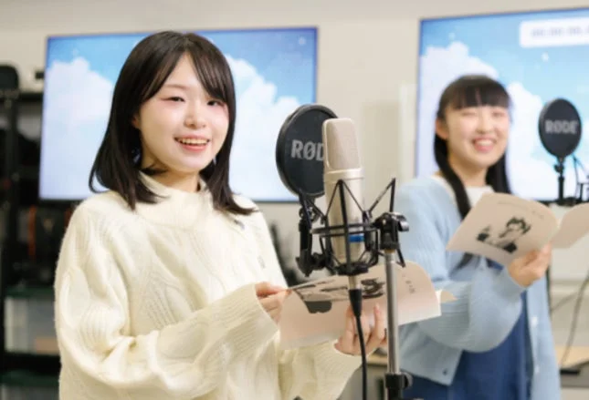 レコーディングスタジオで笑顔でマイクに向かう2人の若い女性。