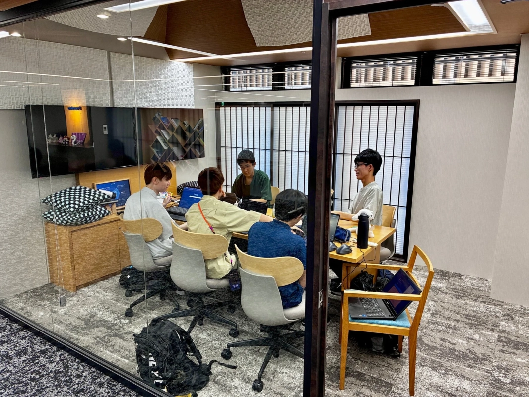 モダンな会議室で5人の若者がノートパソコンに向かい、共同で作業や勉強をしている様子。集中した雰囲気で、活発な議論が交わされているようだ。