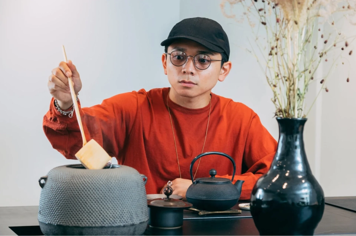 黒いキャップと丸眼鏡をかけた男性が、オレンジ色のシャツを着て、大きな灰色の鍋に木製の道具で何かを入れている様子