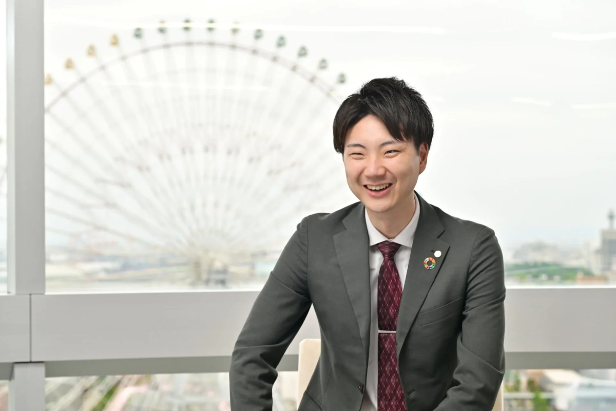 ビジネススーツを着た男性が、窓越しに観覧車が見える室内で笑顔を見せている