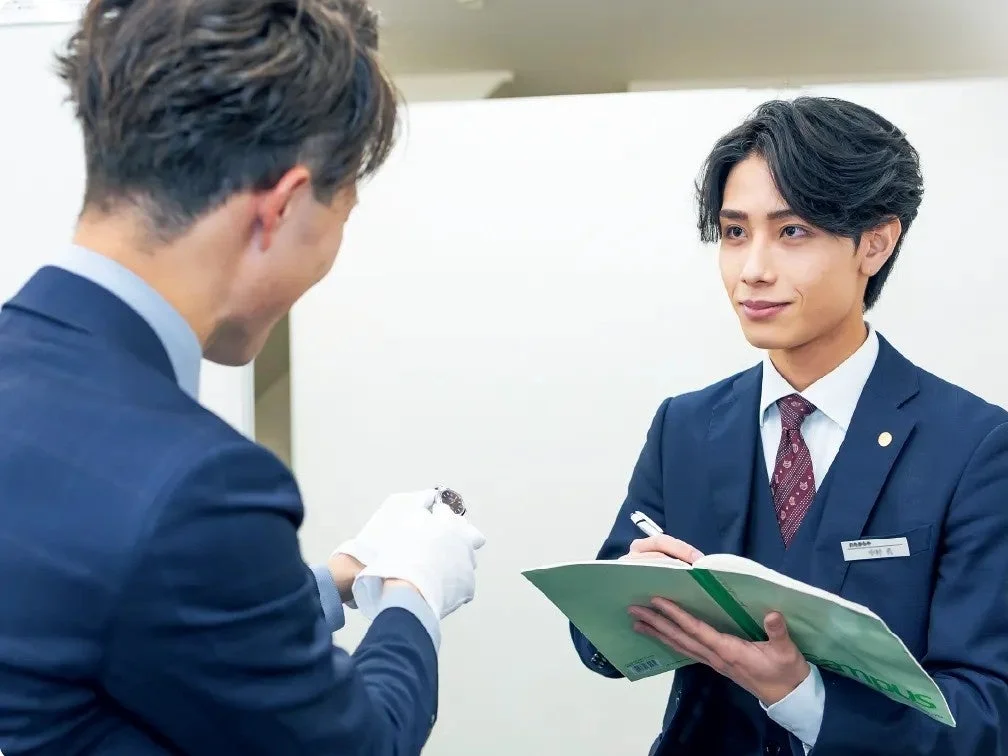 スーツを着た二人の男性が写っており、一人は白い手袋をはめて小さな品物を差し出し、もう一人はノートにメモを取っている