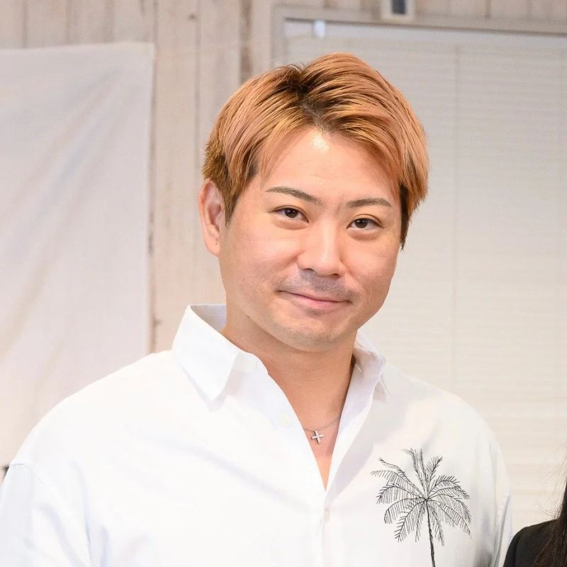 明るい茶色の髪をした日本人男性が白いシャツを着て、カメラに向かって穏やかに微笑んでいます